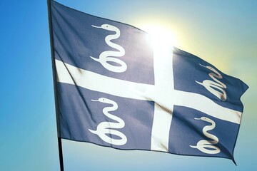 Martinique territorial flag waving in sunlight close-up &ndash; French overseas region banner blue field white cross with four white snakes patriotic Caribbean island symbol background blue sky sunny day
