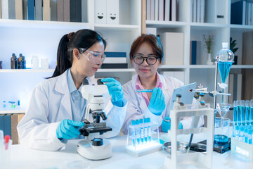 Two female scientists in white lab coats working together in a bright laboratory with microscopes,...
