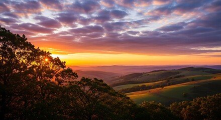 Golden hour sunset paints a tranquil rolling landscape. Sky bursts with vibrant oranges, purples, yellows. Picturesque evening vista over hills, silhouetted trees