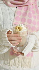 Woman in white sweater and pink scarf holding cup of cream coffee. Concept of wellness and lifestyle branding, illustrating comfort, seasonal rituals, and cafe culture at winter resorts.