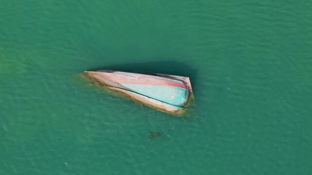 Static overhead aerial view of capsized colorful fishing vessel partially submerged in vivid turquoise bay with shadow visible