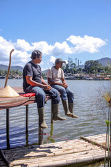 Two Fishermen Relax on Countryside Pier Overlooking Tranquil Lake with Village in the Background