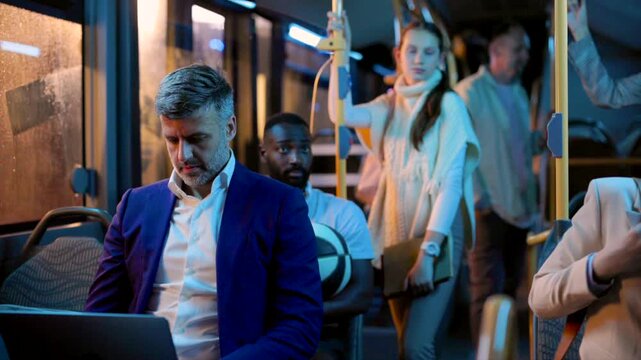 Busy middle aged businessman working on his laptop while commuting home on a crowded bus at night, surrounded by other passengers and looking out the window as the vehicle stops