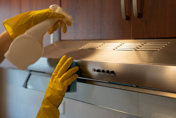Woman in gloves cleaning and wiping stainless steel oven hood in modern kitchen. Hygiene, sanitation, and housekeeping.