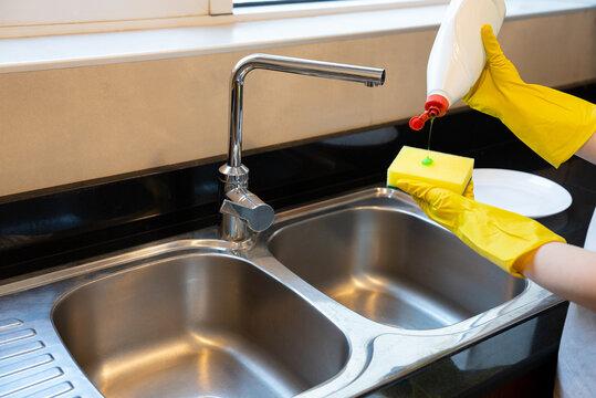 A hand squeezes dish soap onto a sponge above the sink, preparation for washing, advertising-ready for cleaning products and household routine.