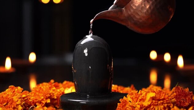 A black Shiva Lingam is being anointed with water from a copper pot. Surrounding it are orange marigold flowers and lit candles, creating a serene atmosphere.