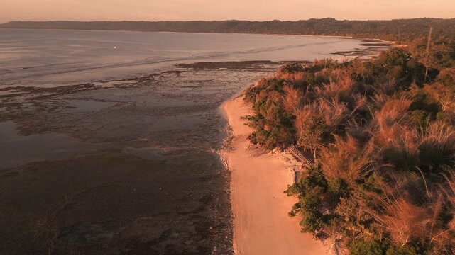 Aerial perspective captures a peaceful coastal scene at sunset, highlighting vibrant greenery and sandy beach, with a gentle drone pan enhancing the serene atmosphere