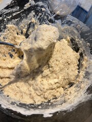 Thick, sticky bread dough with visible grains being mixed in a glass bowl using a metal spoon