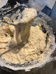 Thick, sticky bread dough with visible grains being mixed in a glass bowl using a metal spoon