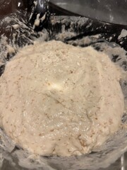 Thick, sticky bread dough with visible grains being mixed in a glass bowl
