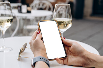 Mockup woman's hand holding smartphone with a blank screen at a chic table, accompanied by a glass of white wine and sunglasses, ideal for mock up use