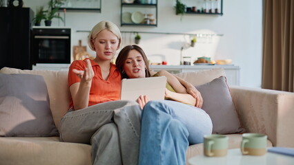 Lesbians looking tablet sitting couch living room morning. Lgbt couple online