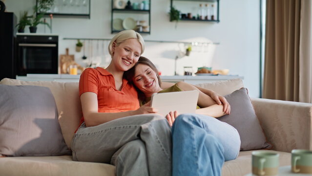 Cuddling girls looking tablet sitting sofa cozy home. Smiling women browsing pad