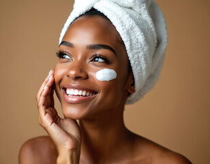 Smiling young african american woman with towel on head applies white cream on cheek. She has clear smooth skin and looks happy enjoying facial treatment.