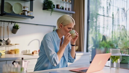 Blonde student typing laptop sipping coffee at light morning kitchen closeup