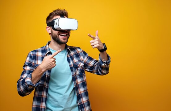 Young man laughs with VR glasses. Wears smartwatch, plaid shirt. Points to empty space, makes gun gesture. Enjoys virtual experience on yellow background. Modern fun. Guy explores new digital world