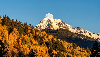 Snow-capped mountain with full moon above peak, surrounded by autumn forest in vibrant orange and yellow under clear blue sky for editorial nature photography seasonal decor and poetic alignment-theme