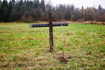 A charred wooden cross and a shovel stand stark in a desolate autumn field. Overcast skies loom over the eerie, rural scene, perfect for Halloween vibes