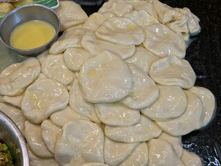 A close-up pile of numerous thin, flattened Roti dough circles, likely buttered or oiled and ready to be cooked, with a small silver bowl of liquid shortening or butter nearby.