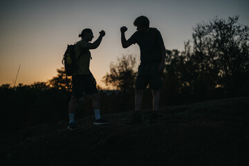 Silhouetted hikers share a fist bump on a trail at sunset, embodying friendship, fitness, and...