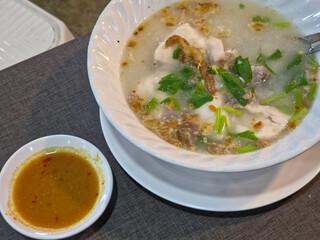 Overhead close-up of a bowl of fish rice porridge garnished with herbs and fried garlic, served with a chili dipping sauce on a dark gray table.