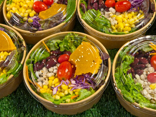 Close-up of multiple ready-to-eat mixed salads in round paper bowls with clear lids, featuring greens, corn, beans, cherry tomatoes, and a slice of pumpkin or squash.