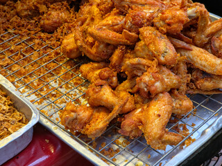 Pile of crispy, golden-brown fried chicken wings and pieces on a wire cooling rack, with loose fried batter and a tray of fried shallots or garlic nearby, suggesting a food stall or market setting.