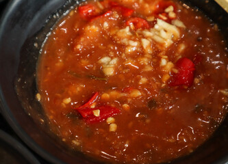 A close-up, high-angle shot of a dark bowl filled with a thick, orange-red suki sauce. The sauce is chunky, featuring visible pieces of red chili and minced white garlic