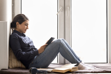Smiling young female student using phone sitting at home windowsill
