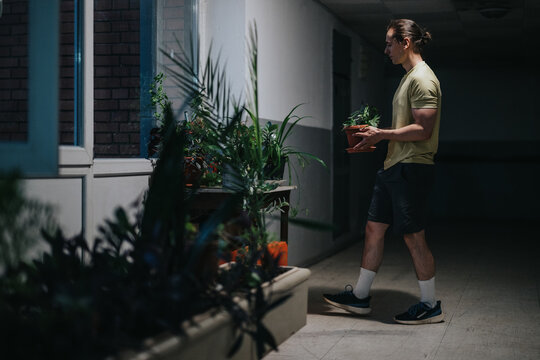 A man in a beige shirt and shorts walks along a corridor, carrying a small potted plant while a row of indoor plants lines the windowsill, creating a calm, urban gardening scene. - Powered by Adobe