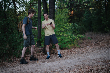 Fototapeta premium Two young men with backpacks stand on a dirt trail in a green forest, one showing something on his phone while the other looks on. They appear to plan or share directions during a hike.