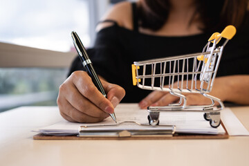 Woman signing financial document with a shopping cart model symbolizing online shopping, e-commerce planning, loan agreement, retail finance, and investment decision. © Lahmz
