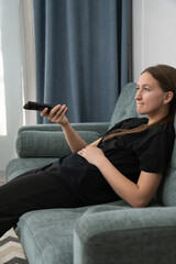 Obraz premium Comfortable young woman lounging on a sofa, watching television and holding a remote control, savoring a peaceful day at home filled with relaxation and leisure. Vertical photo