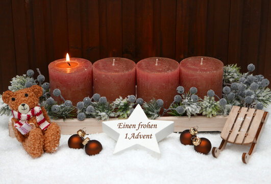 Erster Advent: Rostfarbene Adventskerzen geschm&uuml;ckt mit Teddyb&auml;r, Weihnachtskugeln,und einem Schlitten im Schnee. Auf einem Stern steht der Text einen frohen 1. Advent.
