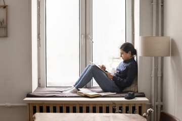 Busy young female remote student using tablet sitting on windowsill