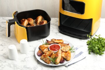 Delicious grilled chicken wings, sauce and spices near air fryer with cooked drumsticks on white marble table