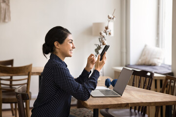 Confident young woman texting message on smartphone by notebook screen