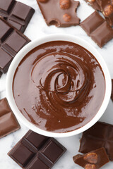 Bowl with melted chocolate and pieces on white marble table, flat lay