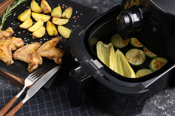 Air fryer with grilled vegetables, chicken wings and potato wedges on grey table, closeup