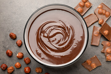 Melted chocolate in bowl, pieces and nuts on grey table, flat lay