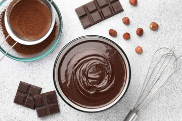 Melted chocolate in bowl, pieces, nuts, cocoa powder and whisk on light grey table, flat lay