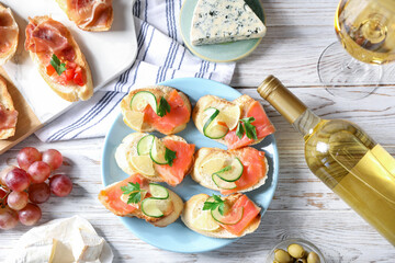 Tasty bruschettas with salmon, cucumber, lemon, prosciutto and wine on white wooden table, flat lay
