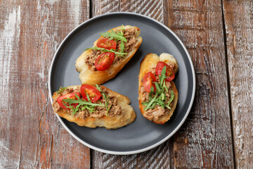 Delicious bruschettas with tuna, tomatoes and arugula on wooden table, top view