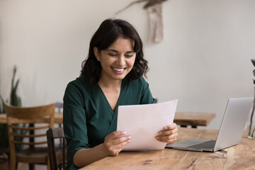 Happy smiling millennial businesswoman reading papers at home office desk