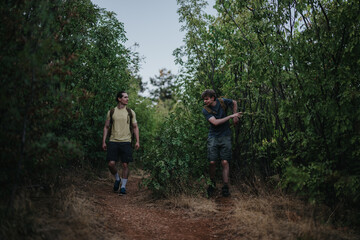 Fototapeta premium Two men hike through dense trees on a dirt forest path, carrying backpacks. They wear casual outdoor gear and share a moment of exploration and friendship in a calm, natural setting.