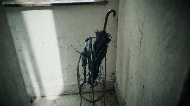 A worn, folded umbrella rests in a rusty metal stand in the corner of a bare, dusty room, bathed in soft daylight.