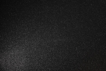 Black glitter texture with fine granular relief, full surface in sharp focus while subtle scattered...