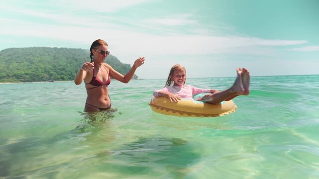 Laughing girl kicks her legs while floating in sea ring, splashing around with mother nearby. Playful summer activity shows emotion, joy, freedom and fun in warm shallow waters.