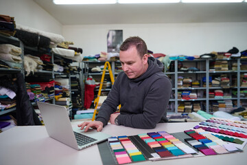 Fashion designer using laptop in textile warehouse surrounded by fabric swatches and rolls,...