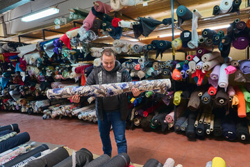 Designer holding roll of printed fabric in warehouse, surrounded by shelves filled with various textiles, showcasing inventory and selection in a textile business.